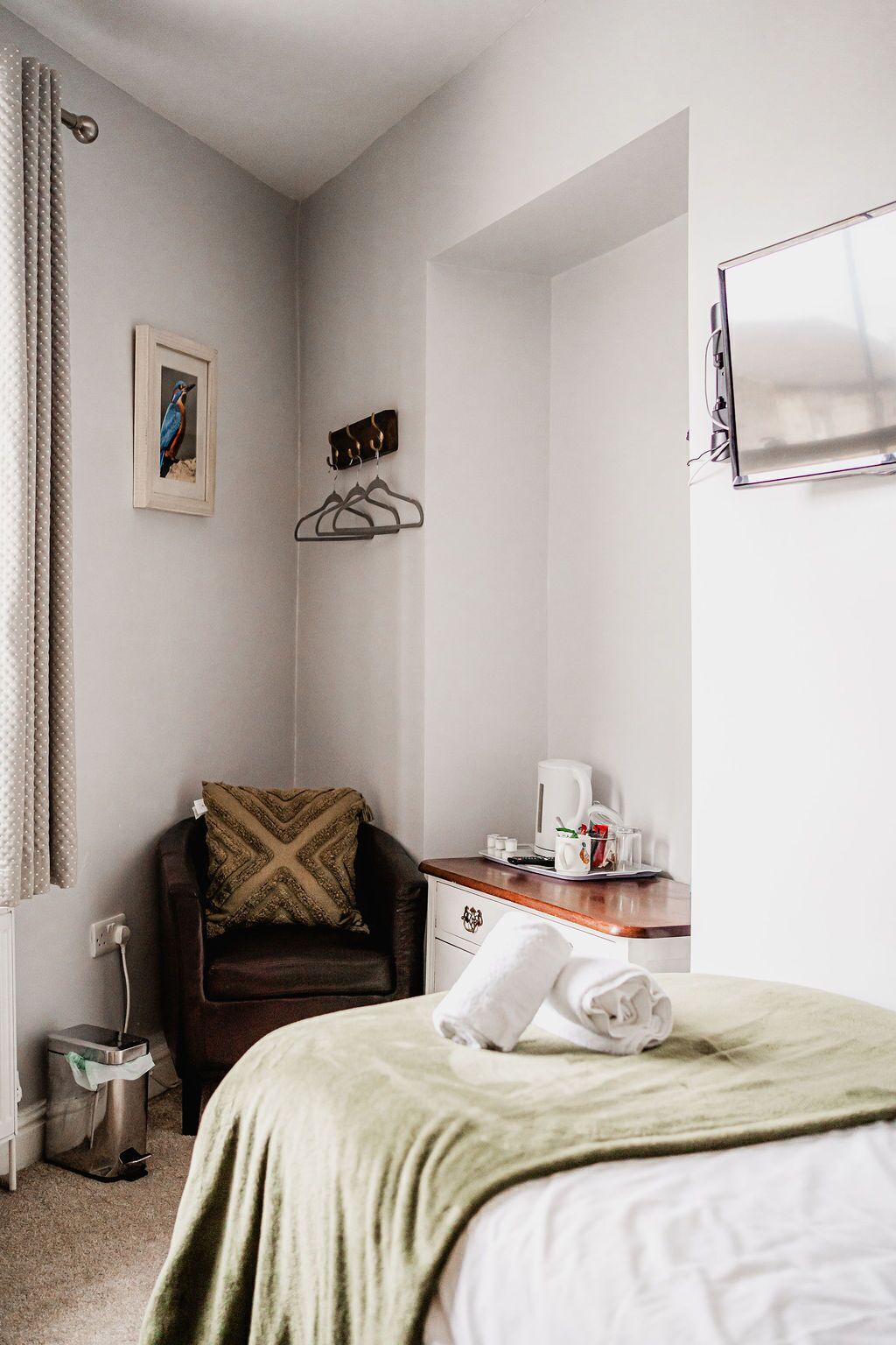 A hotel room with a bed, a chair, a desk with a kettle, and a mirror.