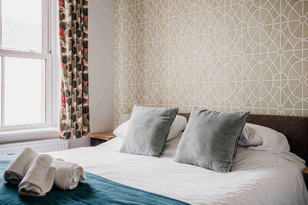 Una cama bien tendida con dos almohadas y una colcha verde azulada, una ventana con cortinas y una pared con un diseño geométrico.