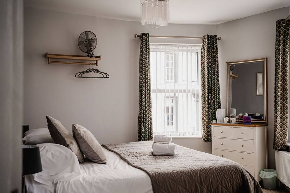 A bedroom with a bed, a bedside table, a mirror, a window with curtains, and a hanging fan.