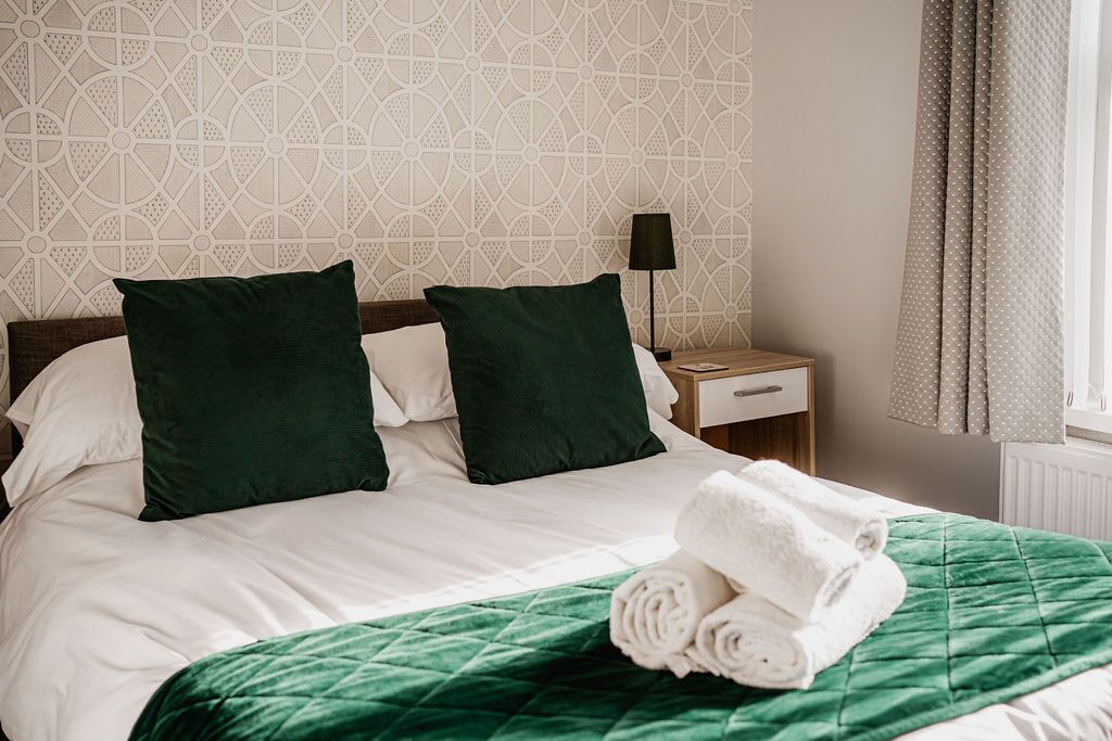 A neatly made bed with white and green bedding, a green throw blanket, and rolled towels on the bed.