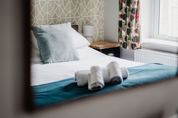 A neatly made bed with rolled towels, a patterned wall, a nightstand with a lamp, and a window with curtains. - The Railway (Skipton)