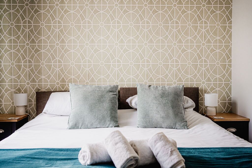 Una cama bien hecha con dos almohadas, una colcha azul y toallas enrolladas sobre un cubrecolchón blanco.