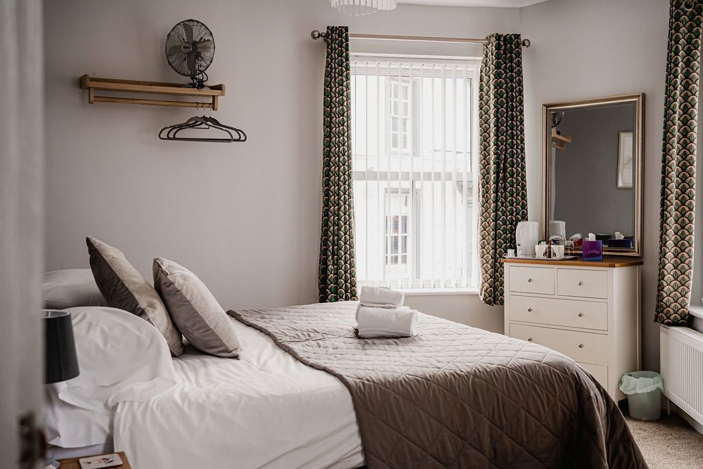 A bedroom with a bed, a bedside table with a mirror, a hanging fan, and a window with curtains.