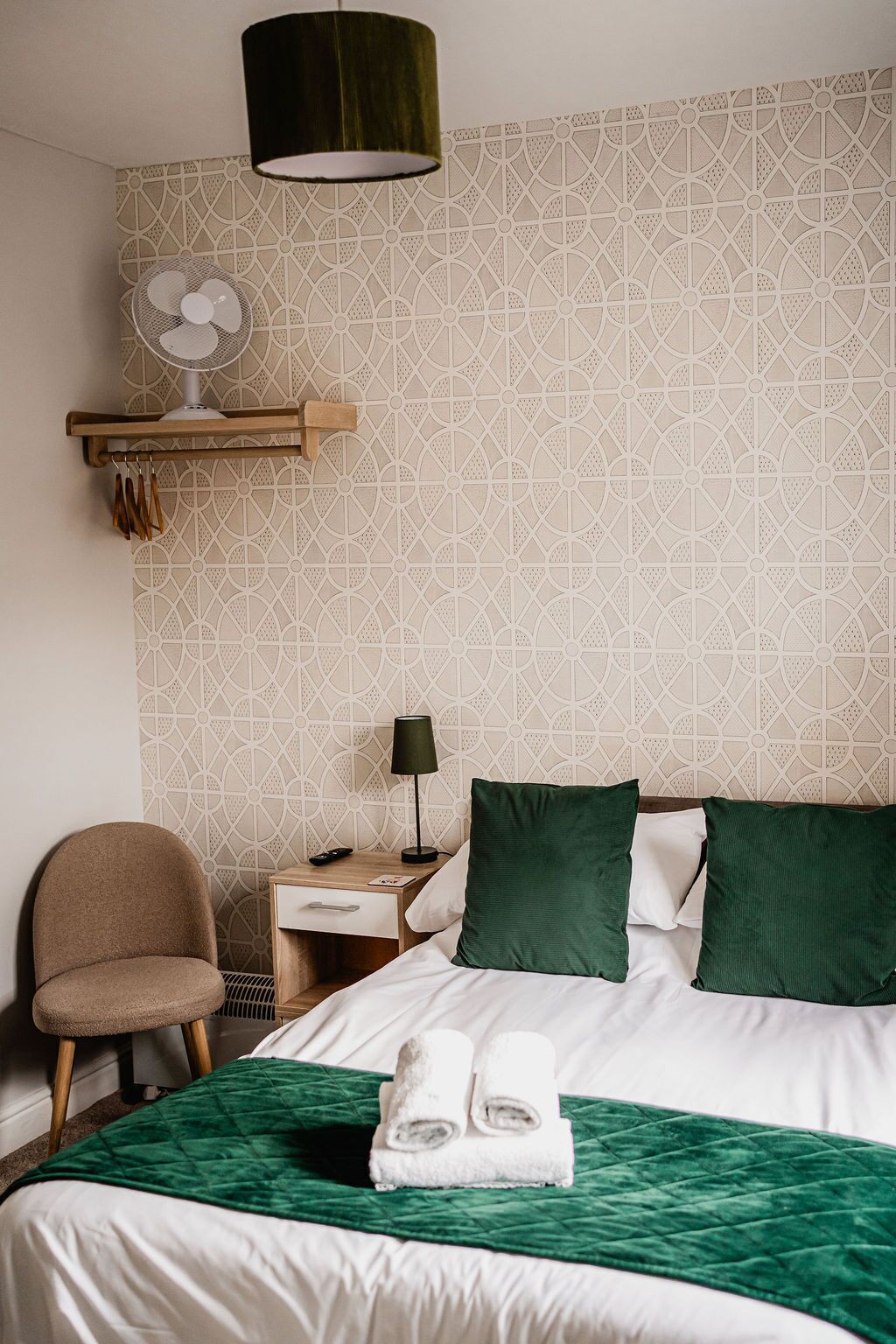 A hotel room with a bed, a chair, a nightstand, a lamp, a wall-mounted fan, and a green quilted bedspread.