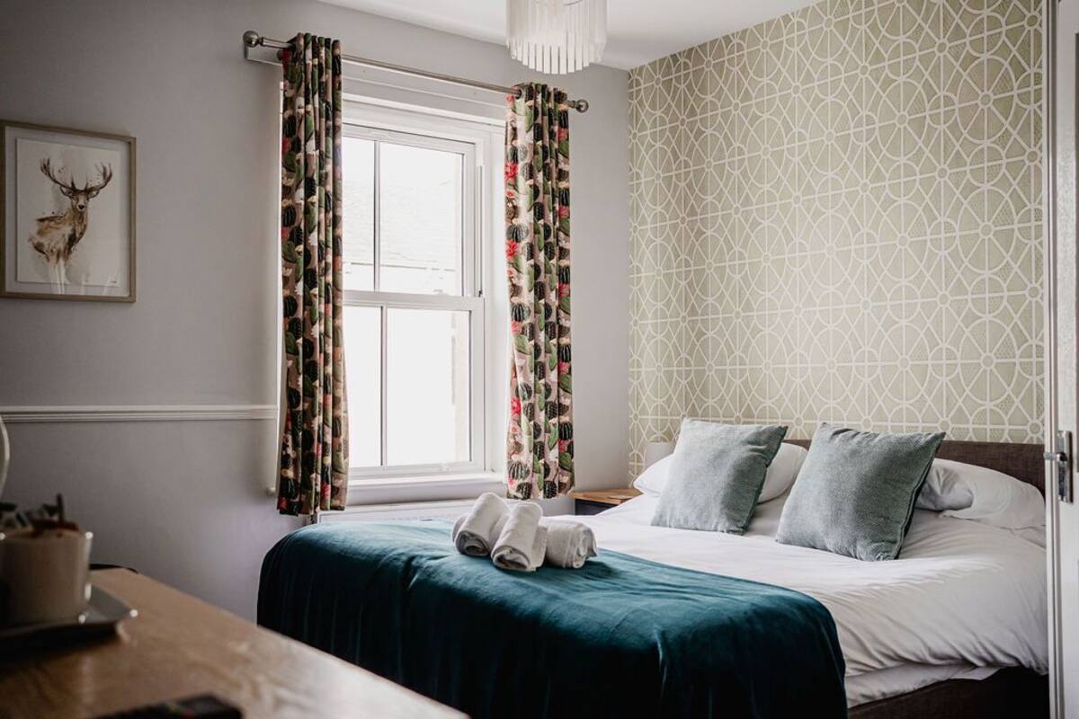 A bedroom with a bed, pillows, a window with curtains, a wall-mounted picture, and a bedside table with a cup and a book.