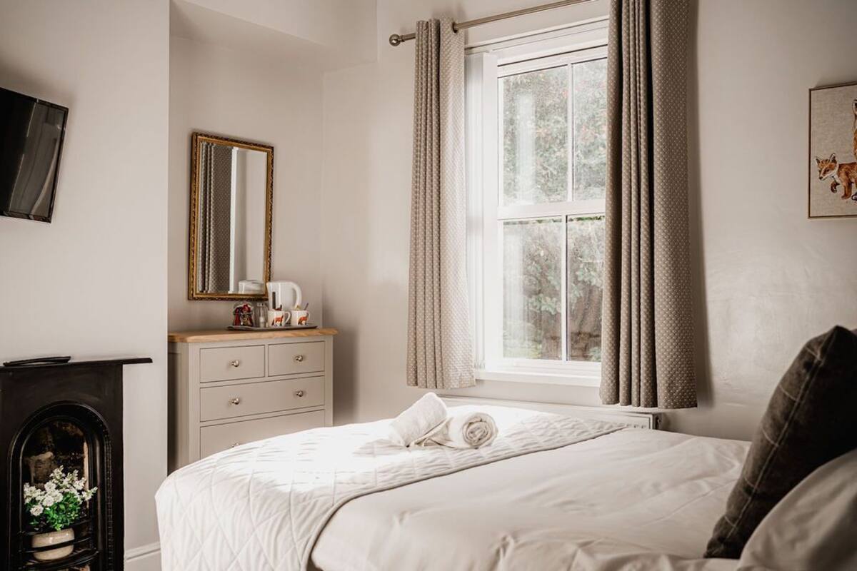 A bedroom with a bed, a dresser, a mirror, a window with curtains, and a fireplace.