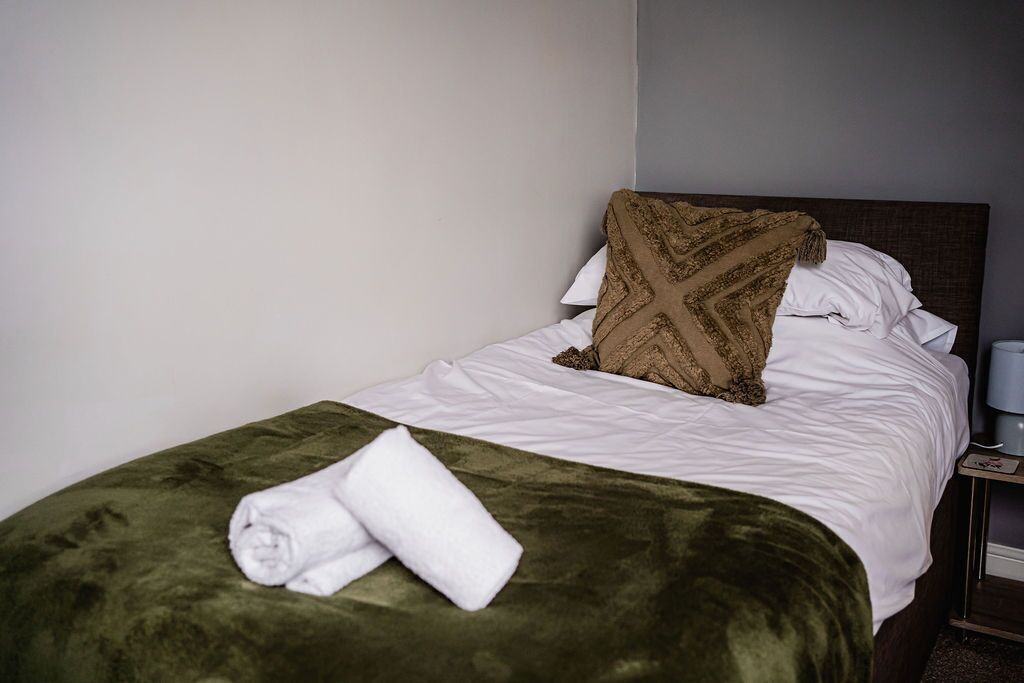 Un lit bien fait, recouvert d’une couverture verte et agrémenté d’un coussin marron, une serviette blanche pliée posée sur le lit, et une table de chevet avec une lampe.
