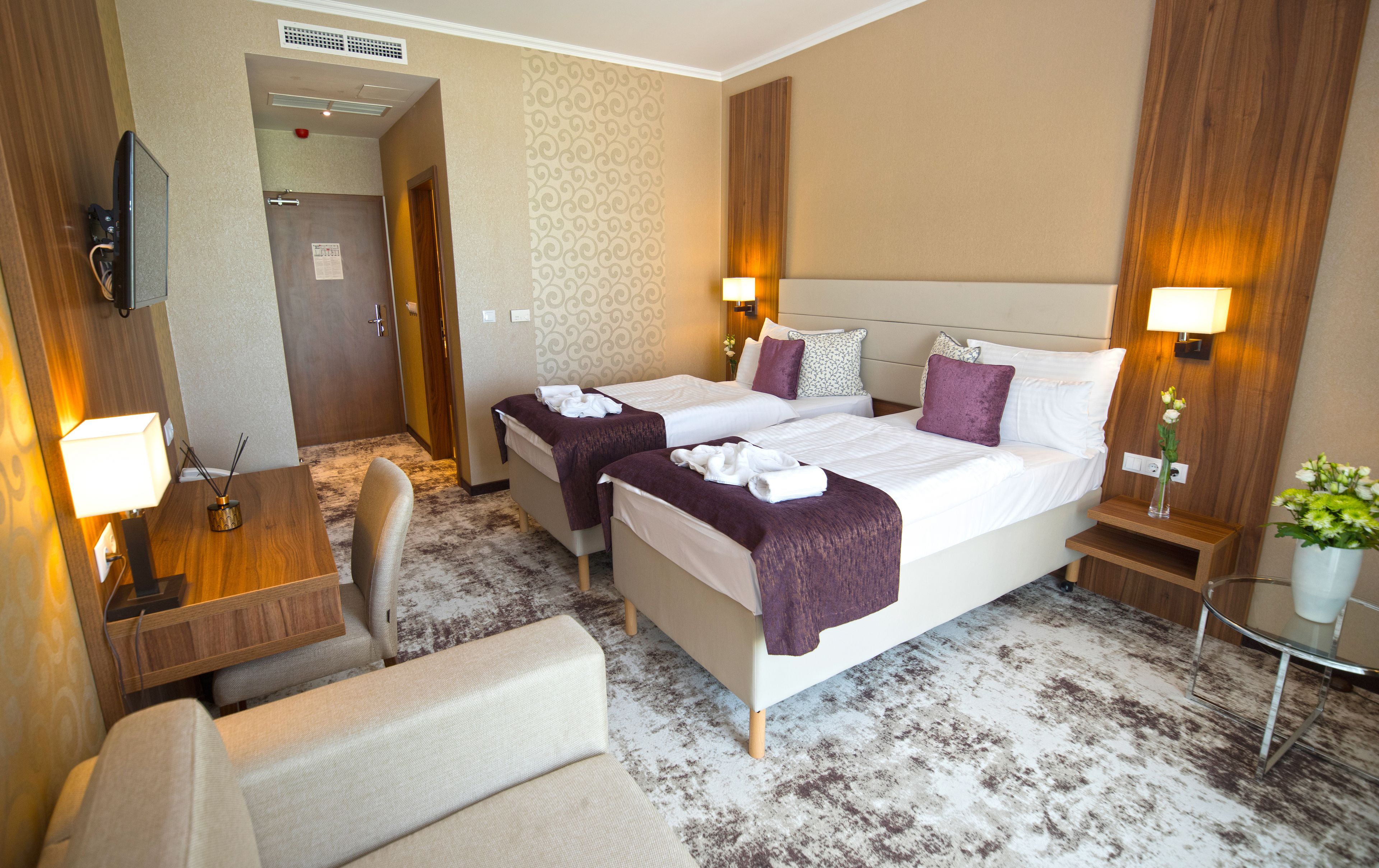 A hotel room with two beds, a desk, a chair, and a small table with a vase of flowers.