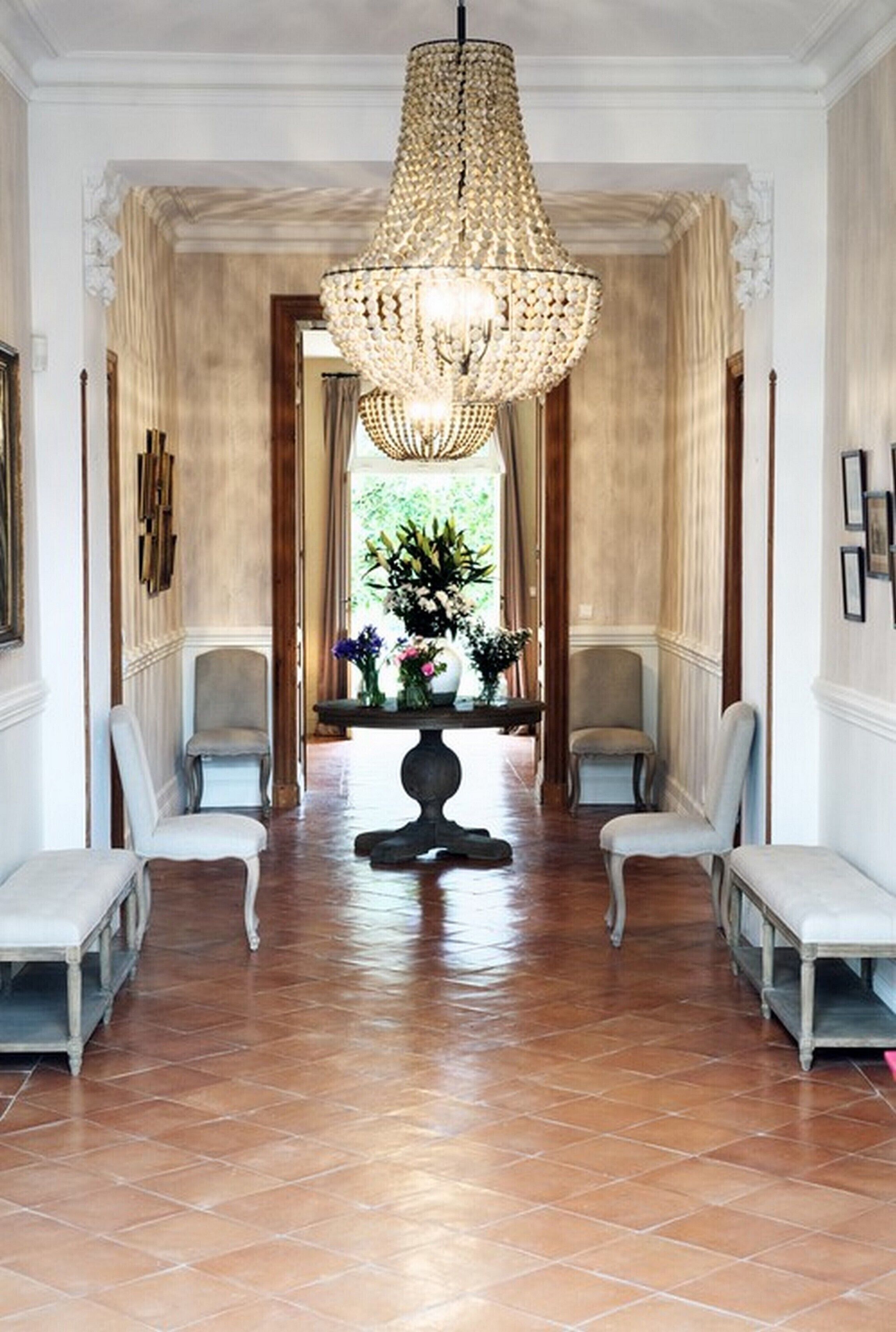 A hallway with a large chandelier, two armchairs, a table with flowers, and a view of greenery through a window.