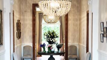 A hallway with a large chandelier, two armchairs, a table with flowers, and a view of greenery through a window.