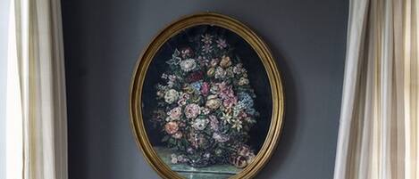 A dining area with a round table, two chairs, a floral painting on the wall, and a window with curtains.