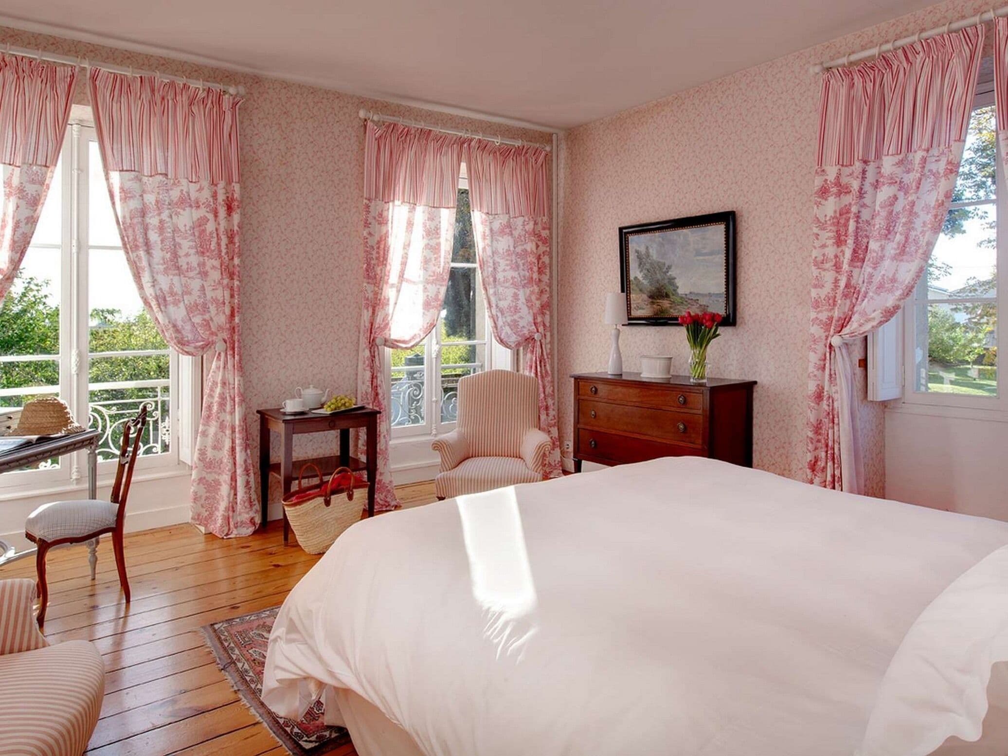 A bedroom with a large bed, a desk, a chair, and a dresser. There is a window with curtains and a view of greenery outside.