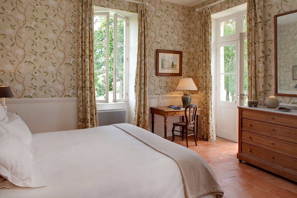 Un dormitorio con cama, escritorio, silla, cómoda y dos ventanas con cortinas.