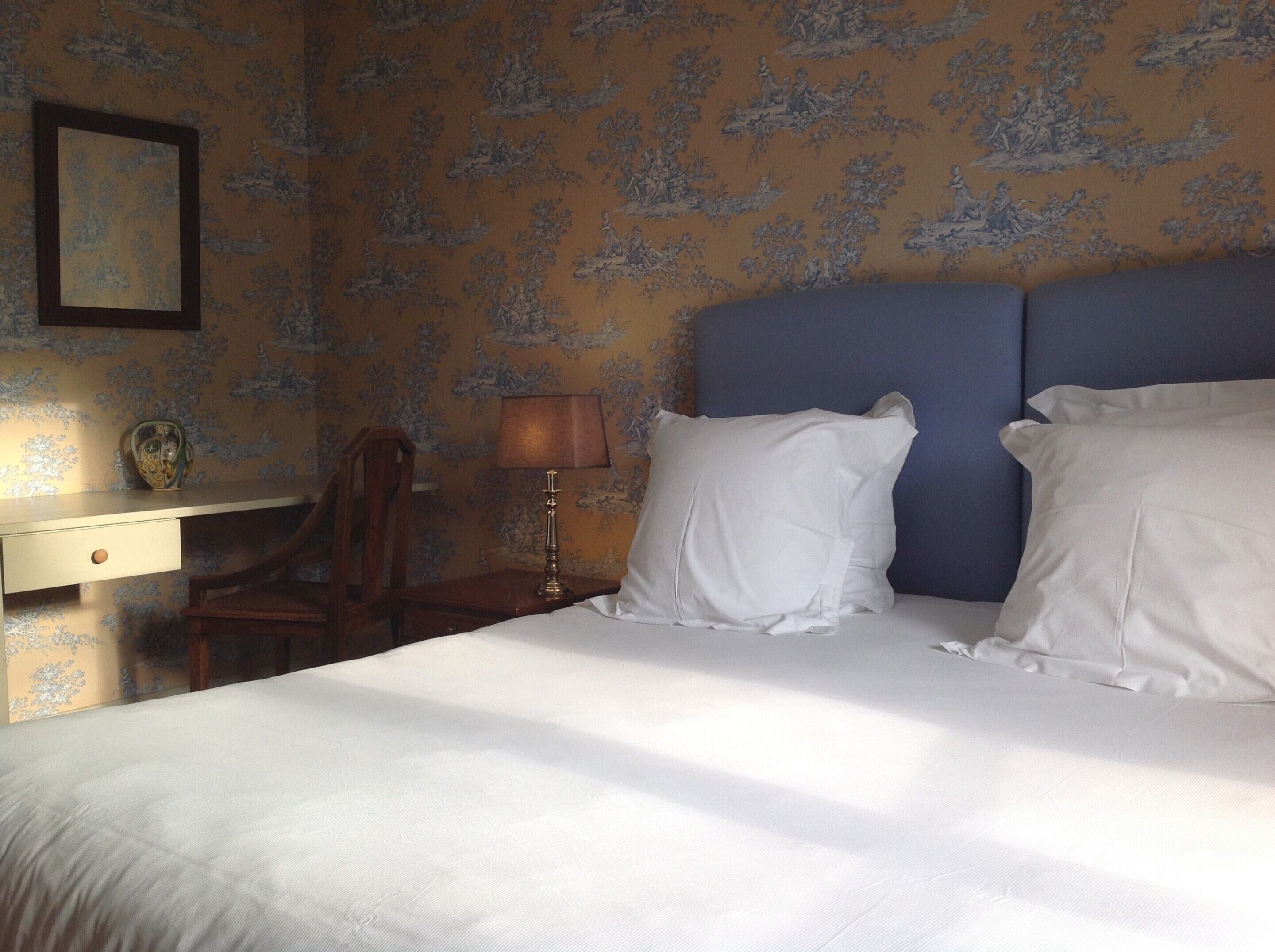 A neatly made bed with white linens, a wooden nightstand with a lamp, and a chair.
