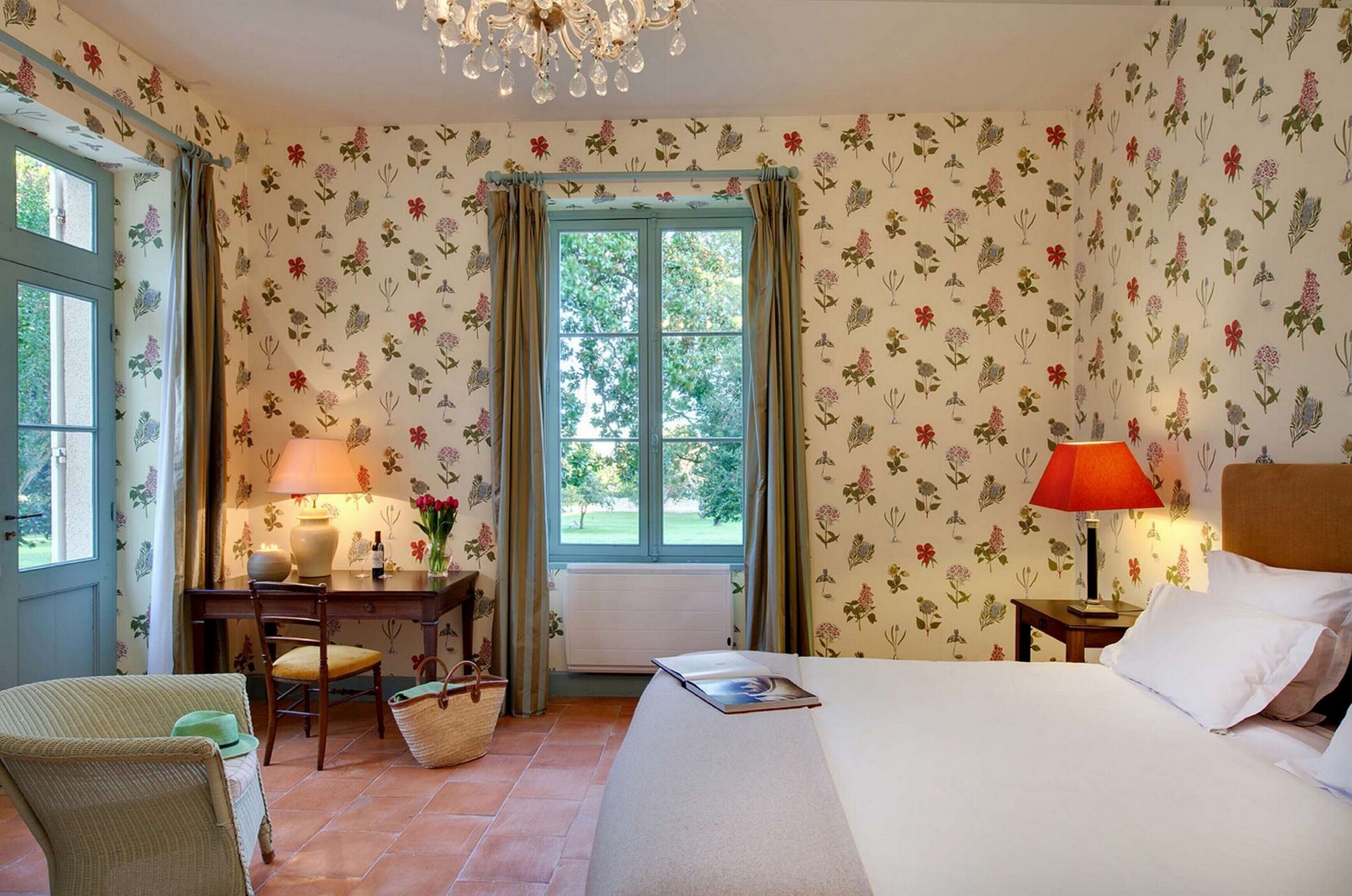 Ein Schlafzimmer mit Blumenmuster-Tapete, einem Bett, einem Schreibtisch mit Lampe, einem Stuhl und einem Fenster mit Vorhängen.