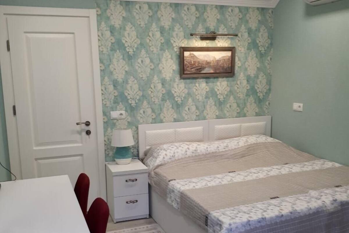 Un dormitorio con cama, mesita de noche, lámpara y un cuadro en la pared.