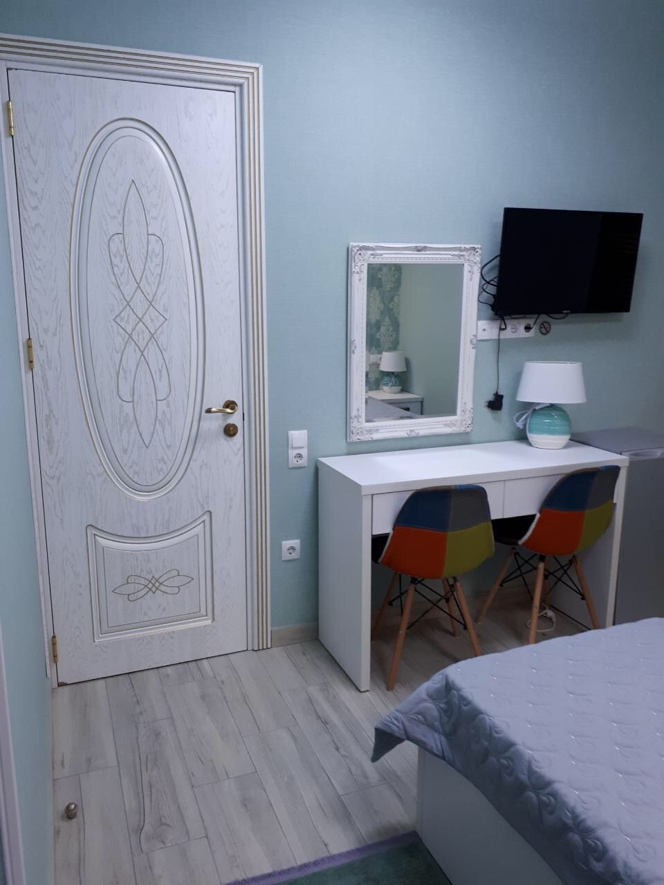 A bedroom with a bed, a desk with a mirror, two chairs, and a television on the wall.