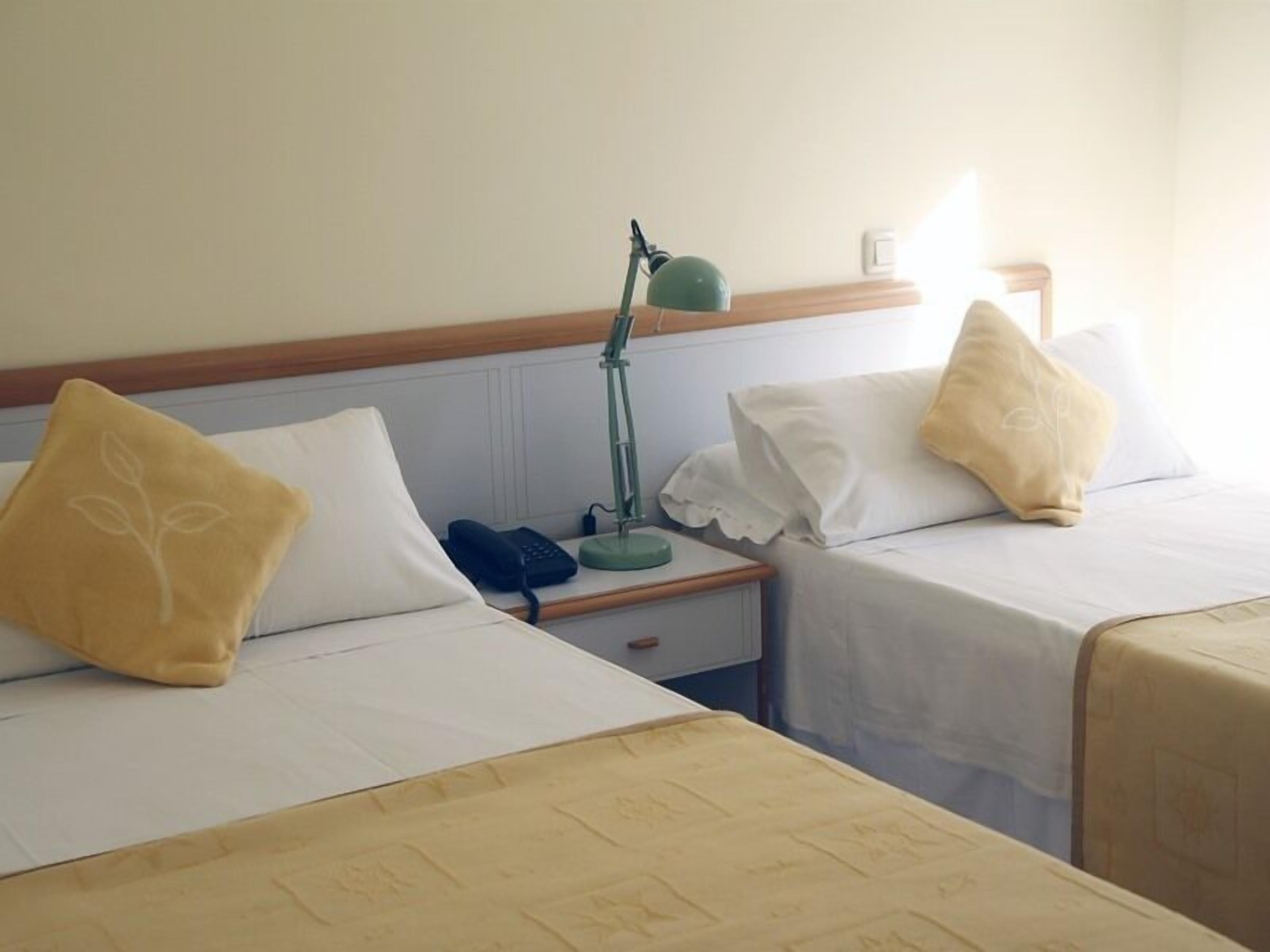 A hotel room with two single beds, each with a pillow and a bedside table.