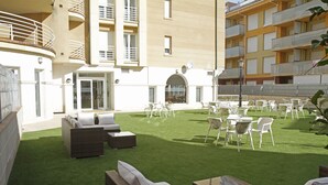 An outdoor patio area with artificial grass, wicker furniture, and white chairs. - Hotel Playa de Merón (San Vicente de la Barquera)