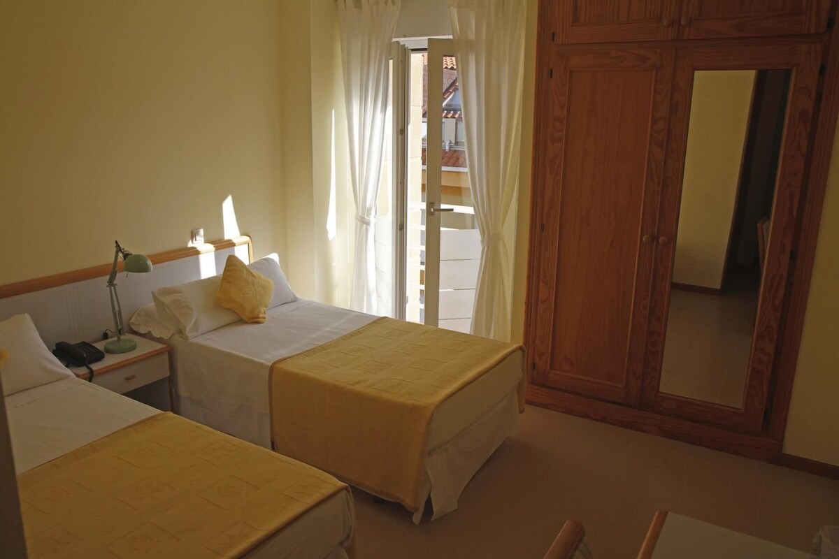 Una habitación de hotel con dos camas, una mesita de noche con lámpara, un armario de madera y una ventana con cortinas.
