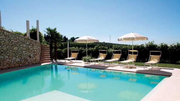 A swimming pool with lounge chairs and umbrellas, surrounded by a stone wall and greenery. - B&B Al Cristo (Sant'Ambrogio di Valpolicella)