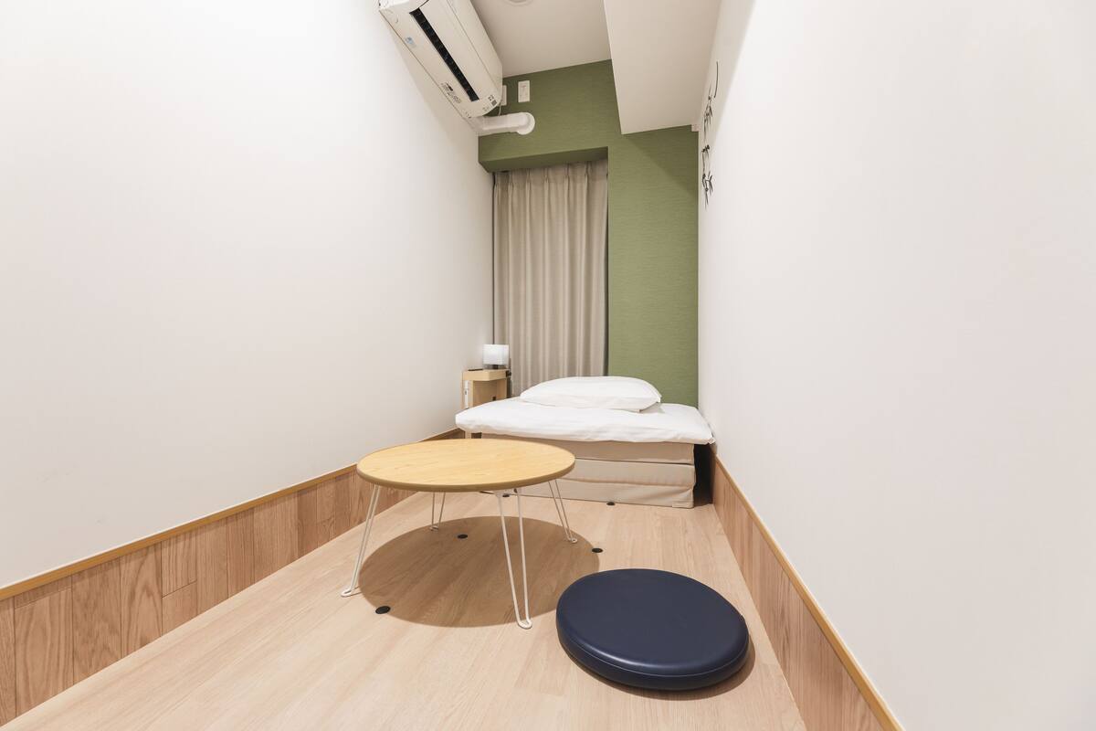 A small, modern room with a bed, a round wooden table, and a small stool.