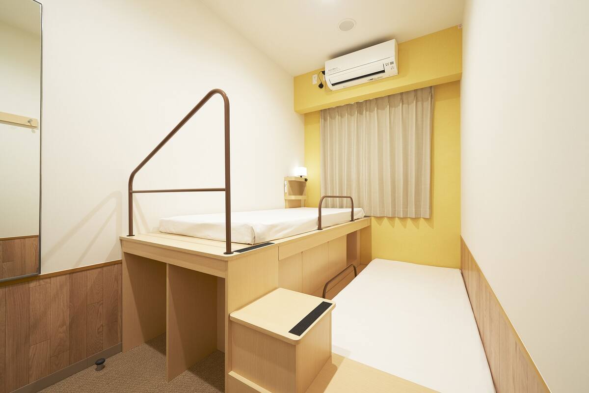 A compact hotel room with a bunk bed, a small desk, a bedside table, a mirror, a window with curtains, and an air conditioning unit.