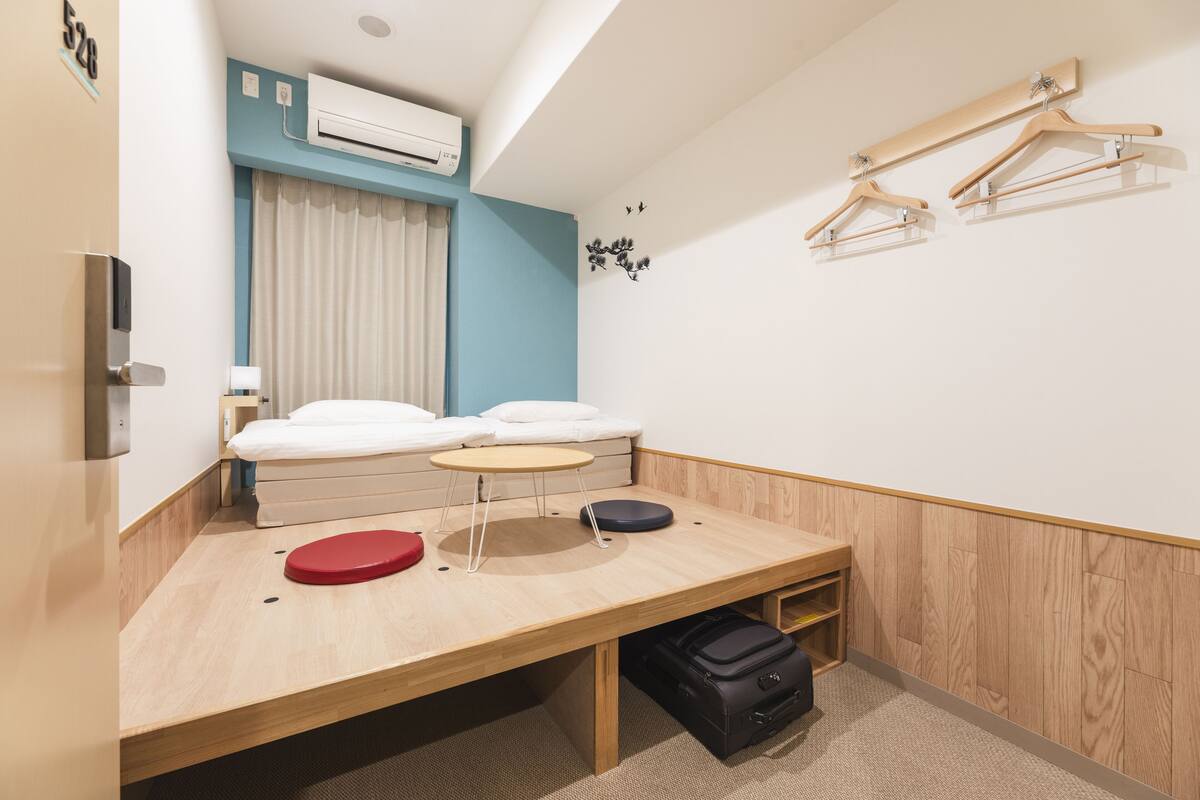 A hotel room with a bed, a desk, a chair, a small table, a red stool, a black suitcase, and a wall-mounted air conditioner.