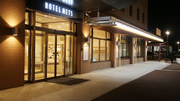 A hotel entrance with a sign reading "HOTEL METS," glass doors, and a covered walkway.