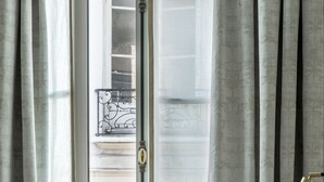 Quarto com uma janela ampla, mesa de cabeceira com abajur e vista para uma varanda com plantas.