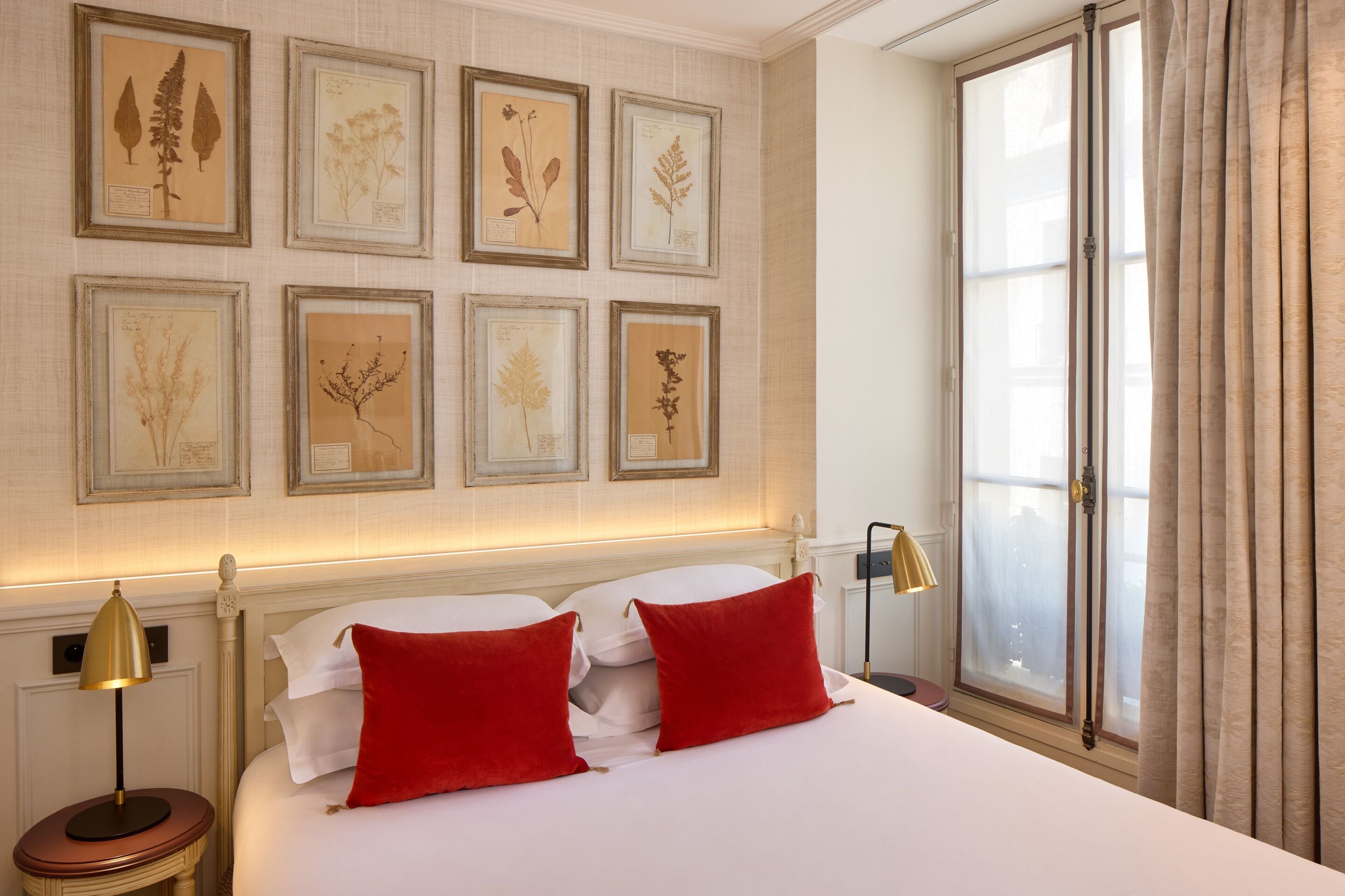 A hotel room with a large bed, red pillows, a nightstand with a lamp, and framed botanical prints on the wall.