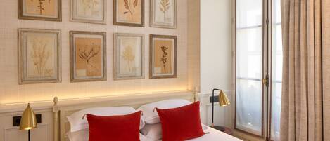 A hotel room with a large bed, red pillows, a nightstand with a lamp, and framed botanical prints on the wall.