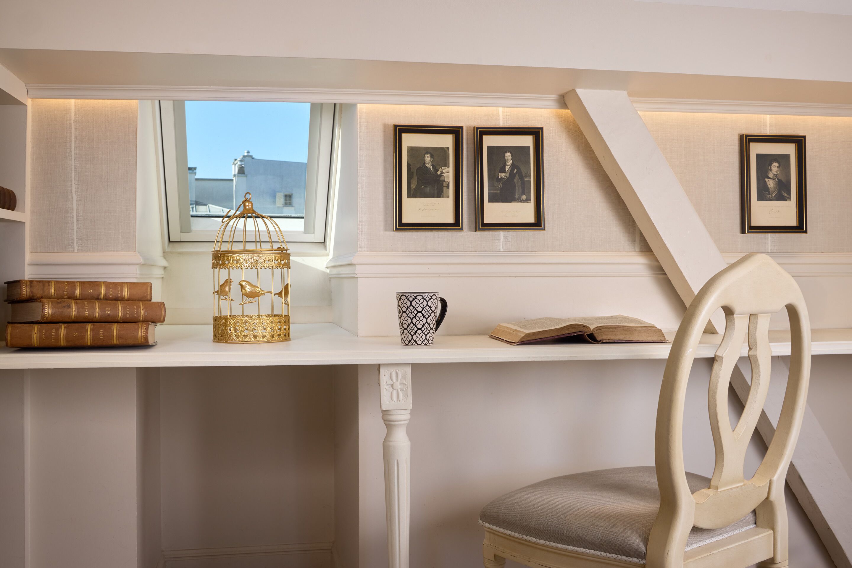Una habitación con escritorio, libros, jaula de pájaros, cuadros enmarcados y una silla.