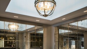 Interior moderno com teto alto, lustre central, área de estar com sofás e poltronas, e janelas amplas refletindo o exterior.