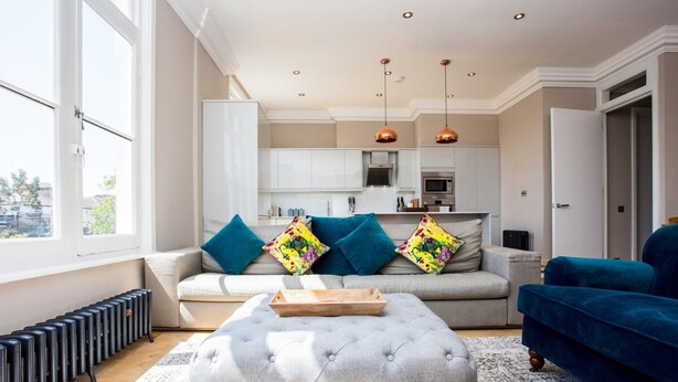 Ein modernes Wohnzimmer mit einem grauen Sofa, einem blauen Samt-Sessel und einem gepolsterten Hocker. Im Hintergrund ist eine Küche mit weißen Schränken und Kupfer-Pendelleuchten zu sehen.