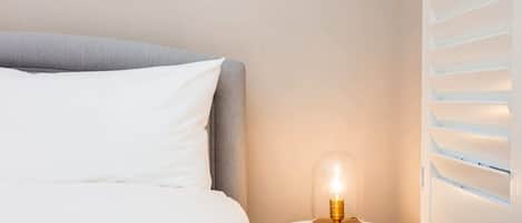 Uma cama bem arrumada com lençóis brancos e cabeceira cinza, um criado-mudo redondo branco com cúpula de vidro transparente, e uma janela com venezianas horizontais.