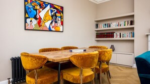 Ein Essbereich mit einem Holztisch und gelben Samtstühlen, einem farbenfrohen abstrakten Gemälde an der Wand und einem Bücherregal mit Büchern und Dekorationsobjekten.