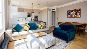Ein modernes Wohnzimmer mit einer Couch, einem Hocker und einem blauen Sessel. Im Hintergrund ist ein Essbereich mit Tisch und Stühlen zu sehen.