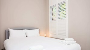 Ein ordentlich bezogenes Bett mit weißen Leinen und zwei aufgerollten Handtüchern, eine Nachttischlampe sowie ein Fenster mit weißen Rollläden.