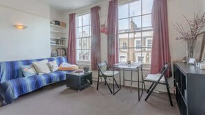 A living room with a sofa, a small table, and a chair. There are bookshelves and a window with curtains.