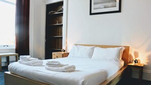 A neatly made bed with white linens and towels, a wooden headboard, a bedside table with a lamp, and a large window with curtains. - Traditional Apartment oer the Meadows (Edinburgh)