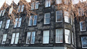 A multi-story building with a dark stone facade, multiple windows, and a row of shops with red awnings at the ground level. - Traditional Apartment oer the Meadows (Edinburgh)