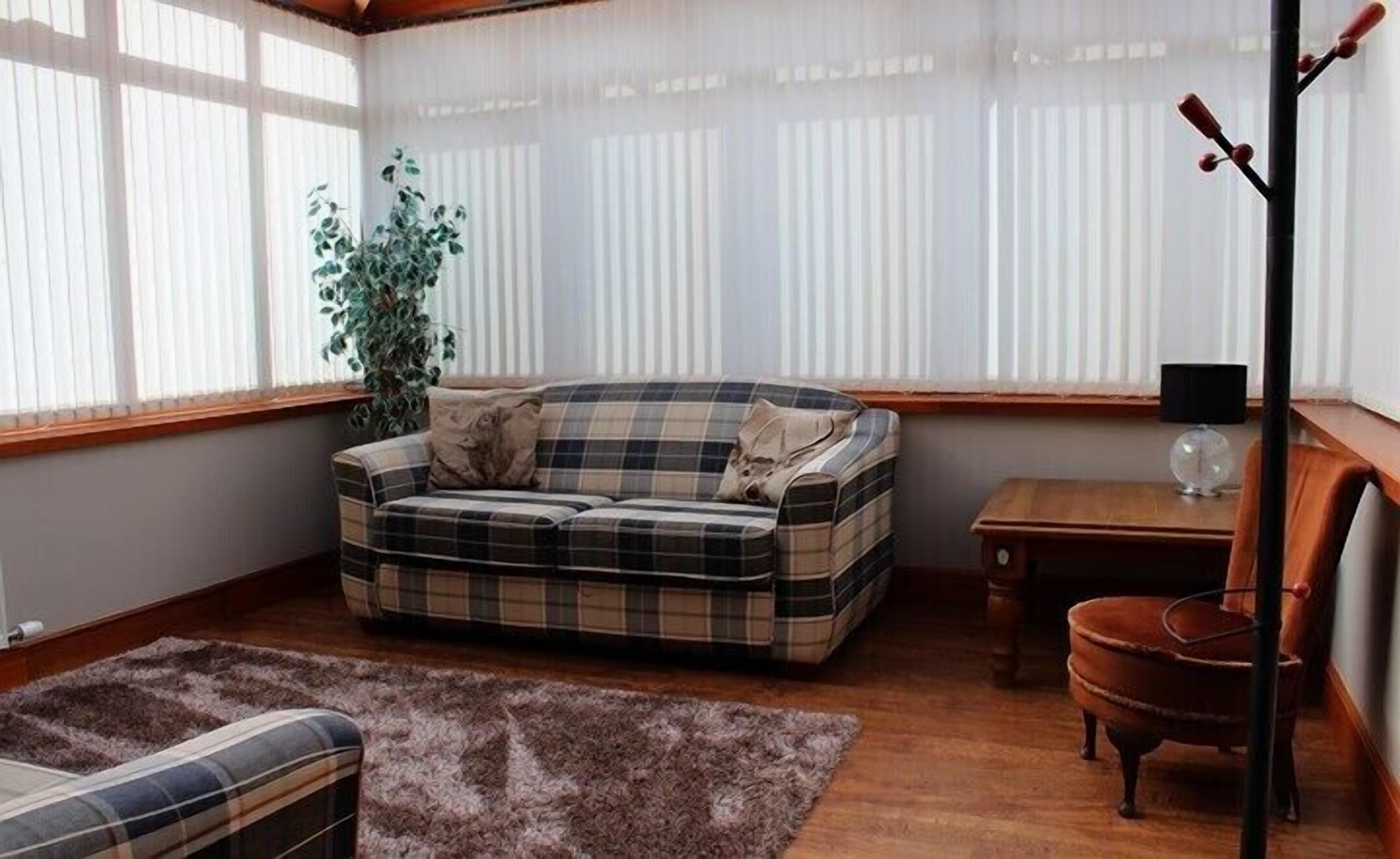 Een gezellige woonkamer met een geruit bankje, een klein houten tafeltje, een stoel en een groot raam met dunne gordijnen.