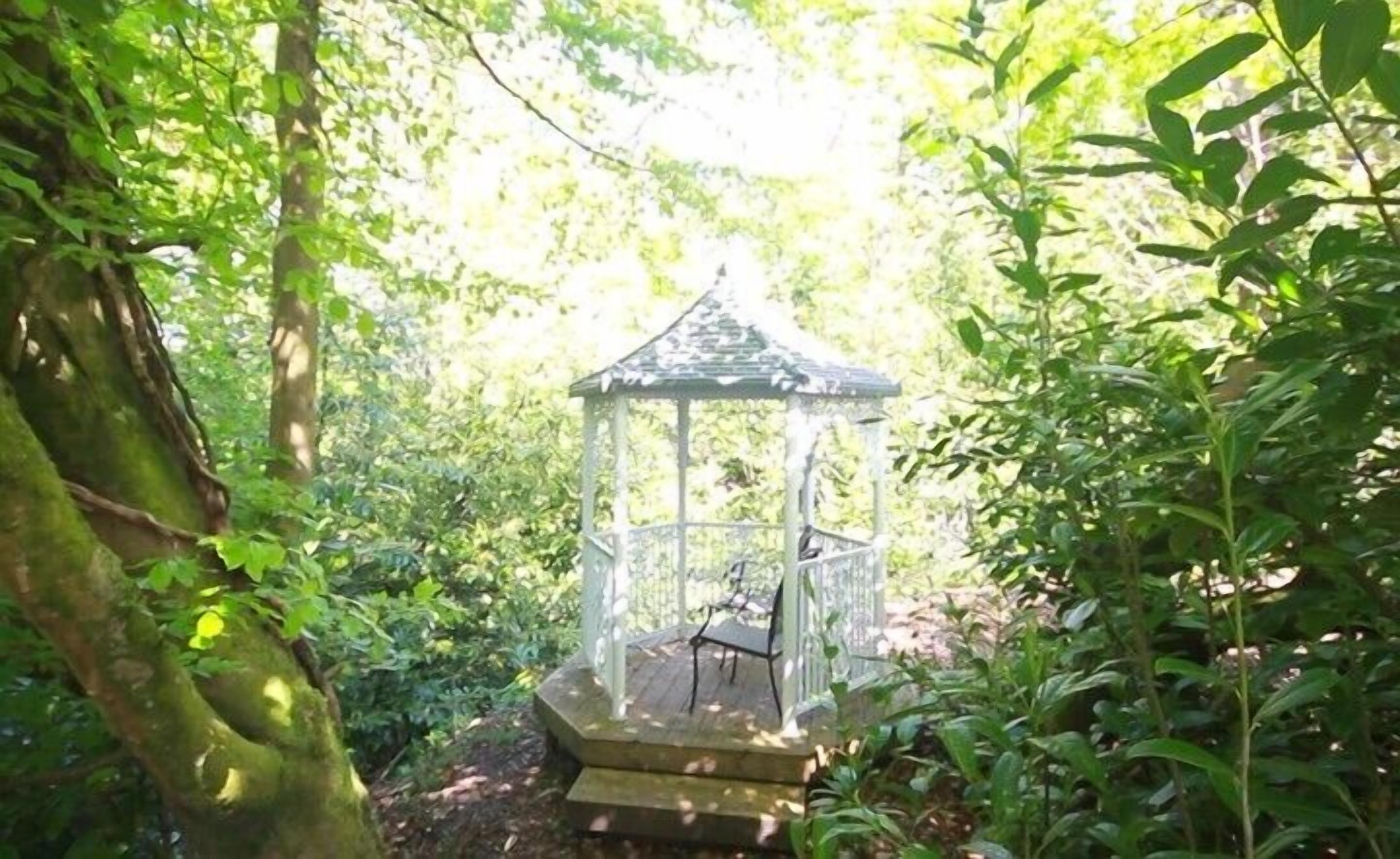 A gazebo with a roof and steps leading up to it, surrounded by dense greenery.