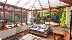 A conservatory with a wooden ceiling and large windows. Inside, there is a plaid sofa with cushions, a small table, and a lamp. - Auchenheglish Lodges - The Smiddy (Alexandria)