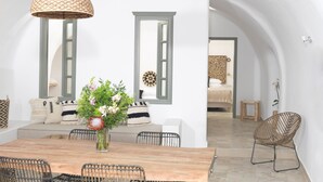 A dining area with a wooden table and chairs, a hanging light fixture, and a view into a bedroom with a bed and a wall-mounted decorative item. - Drops Cave Houses (Santorini)