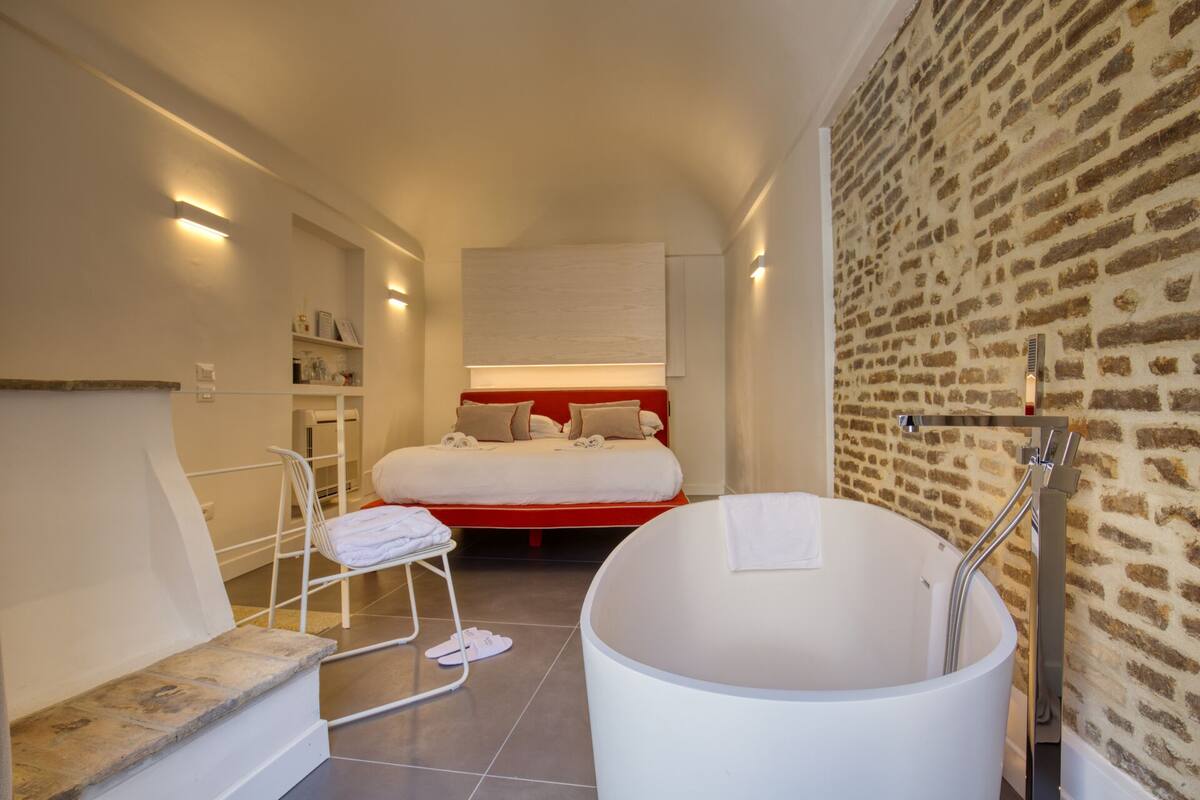 Ein modernes Hotelzimmer mit einer großen Badewanne, einem Bett mit rotem Kopfteil und einer Ziegelwand.