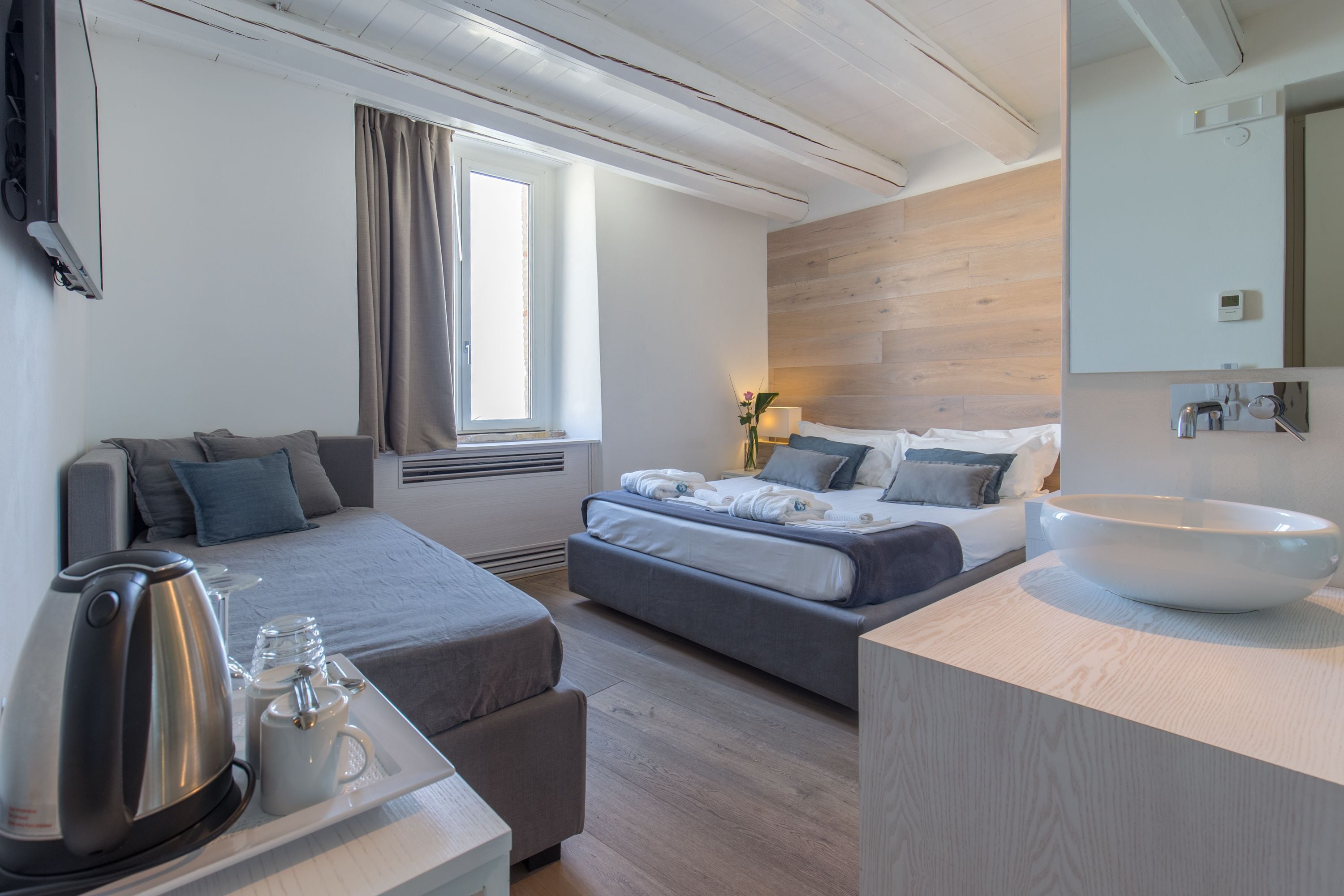 Une chambre d’hôtel moderne avec un grand lit, un canapé, une table basse avec une bouilloire et des verres, un grand bol blanc et un mur en bois.