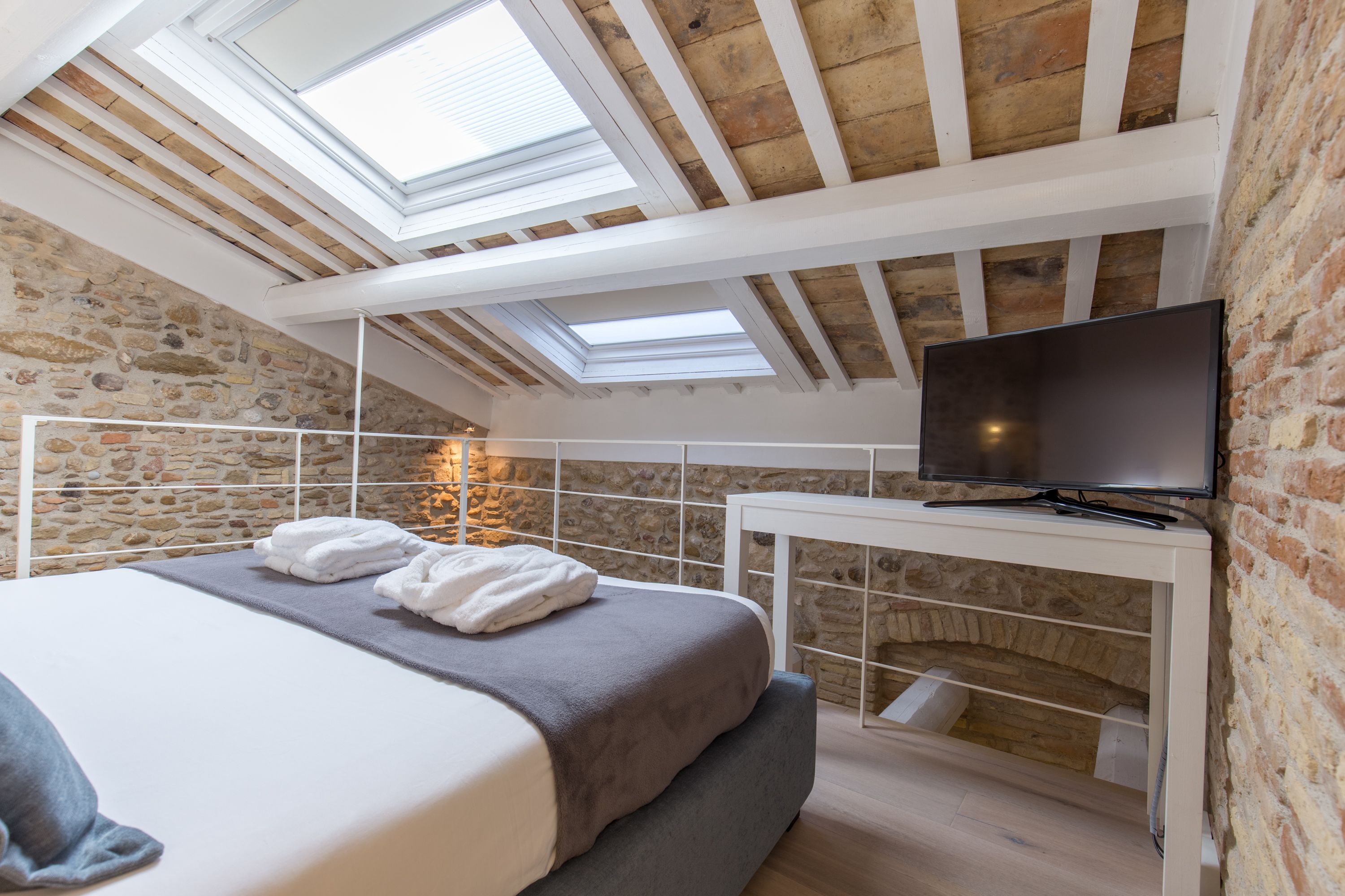 Ein Schlafzimmer mit Steinwand, einem Bett mit grauem Kopfteil, einem Flachbildfernseher auf einem weißen Tisch und Dachfenstern.