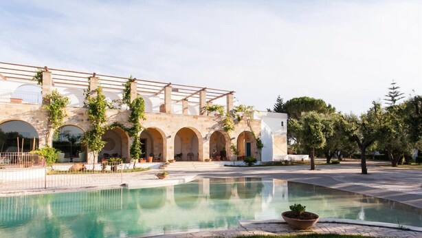 Un área de piscina con una pérgola, arcos de piedra y un edificio blanco.
